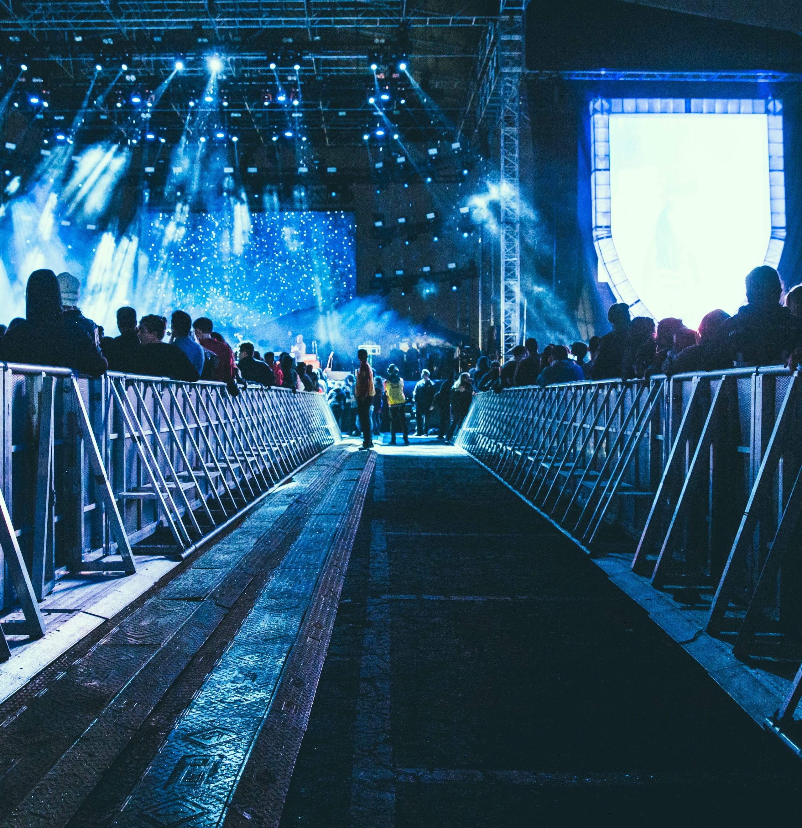 Concertpubliek en verlicht podium, gezien vanaf een blauw verlichte loopbrug